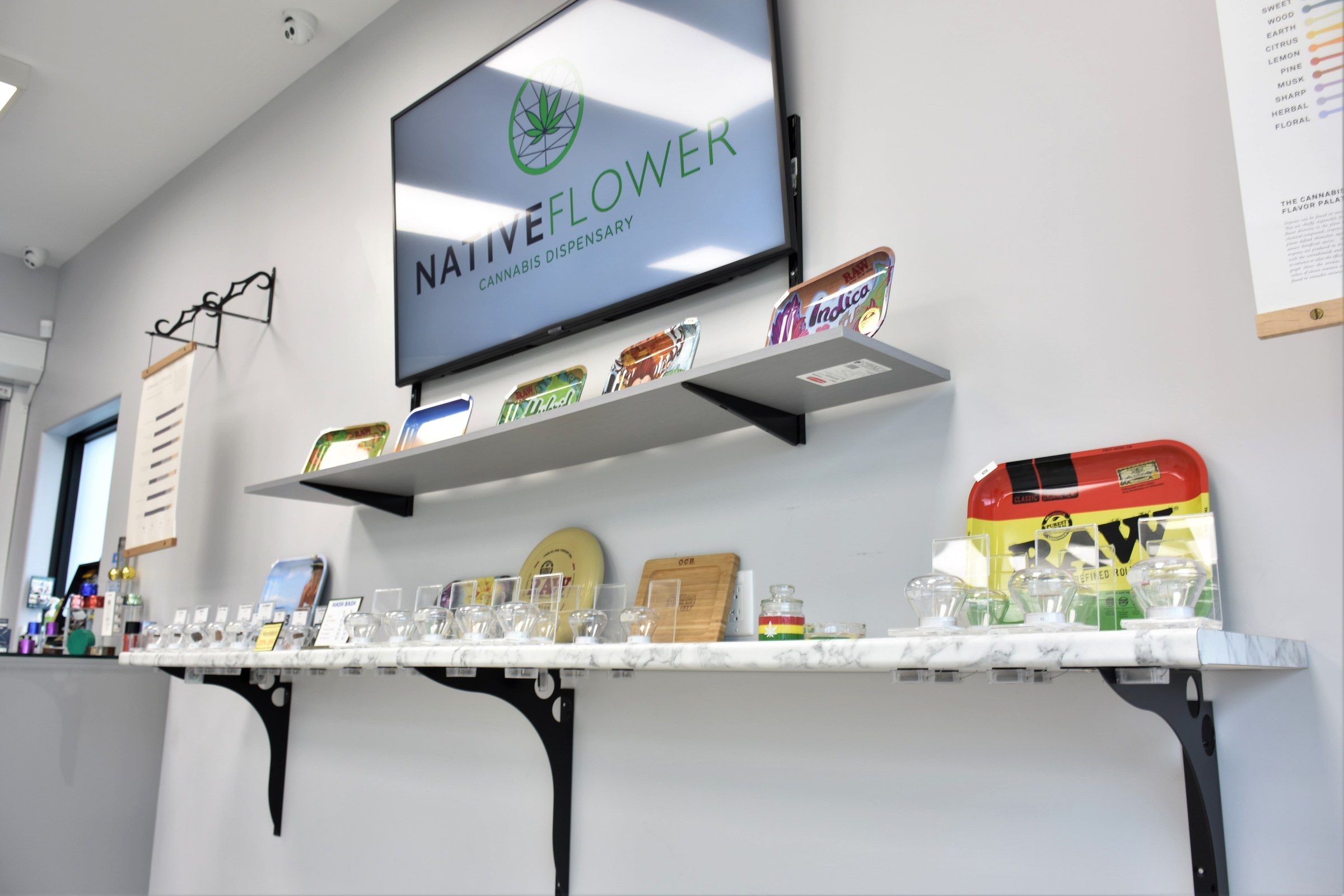 Inside the shop: a wall-mounted screen showing the Native Flower logo above two glass shelves of pipes and glassware, with a marble-topped counter below.